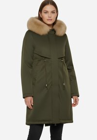 Parka verde oliva con colletto in pelliccia sintetica, chiusura con zip, vita regolabile con cordino e due tasche laterali. Tessuto liscio e design fino al ginocchio.