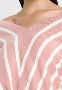 Roze gebreide top met witte horizontale strepen, voorzien van een v-halslijn en een zachte textuur. Focus op het unieke patroon en het kleurcontrast.