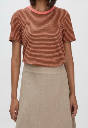 Femme portant un t-shirt à manches courtes rayé marron et rose, rentré dans une jupe beige taille haute, debout devant un fond uni.