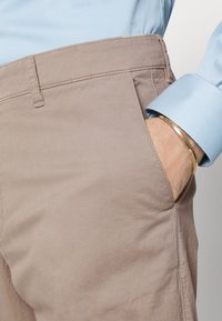 Beige bomullsbyxor med rak passform, med sidofickor och detaljerad sömnad. En del av en blå skjortärm är delvis synlig.