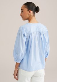 Light blue blouse with puffed sleeves, featuring embroidered details on the cuffs, a round neckline, and a relaxed fit at the back.