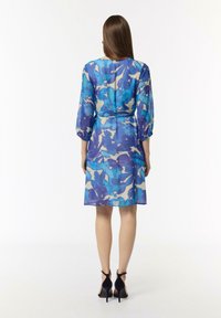 Niebieska i beżowa sukienka w wzory z rękawami o długości trzech czwartych, zaznaczoną talią i długością do kolan. Przezroczysty materiał o luźnym kroju. Czarne sandały na obcasie.