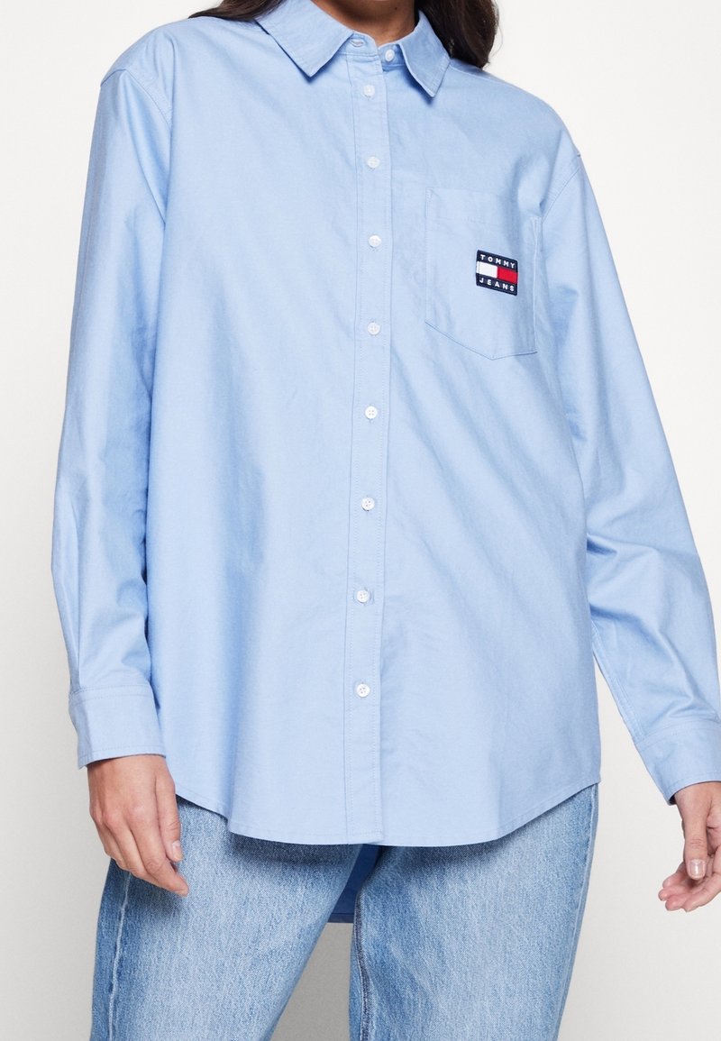 Light blue button-up shirt with a collared neck, long sleeves, and a chest pocket featuring a multi-colored logo patch. Soft cotton texture.