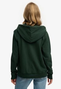 Vrouw met schouderlang blond haar, gekleed in een donkergroene hoodie en lichtblauwe jeans, staand met haar rug naar de camera.