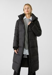 Sort pufferjakke med en lang silhuet, hætte og lynlås. Har quiltede mønstre og ribbede manchetter. Båret over en grå sweater og en hvid skjorte.