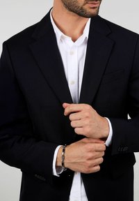 Marinblå blazer med strukturerade axlar, enkel bröstficka, i kombination med en vit skjorta med knappar. Svart accessoar på handleden ger kontrast.