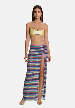 Donna con crop top giallo e gonna lunga a righe multicolori con spacco laterale, abbinati a sandali gialli e orecchini a cerchio d'oro.