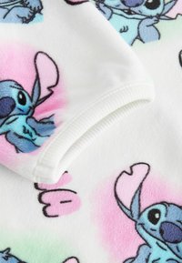 Valkoinen fleece-kangas, jossa on monivärinen sarjakuvahahmo, jossa on sinisiä ja vaaleanpunaisia yksityiskohtia. Käännetty reuna ribbikuvioinnilla ja leikkisällä painatuksella.