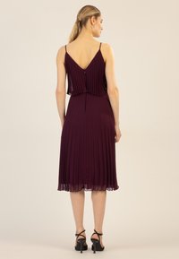 Faltenrock Kleid in Burgunderrot mit einem V-Ausschnitt, Schulterträgern und einem fließenden Saum. Verfügt über einen Reißverschluss auf der Rückseite zur Schließung.