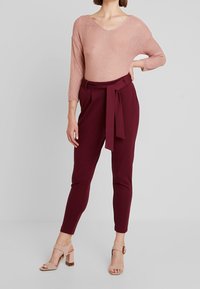 Jasnoróżowy sweter z teksturowanym splotem i dekoltem w kształcie litery V, w zestawie z bordowymi dopasowanymi spodniami związywanym paskiem w talii. Beżowe sandały na obcasie.