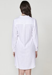 Rochie albă din in, cu mâneci lungi și manșete cu nasturi, având un detaliu la spate și un tiv ușor curbat. Textură fină a țesăturii.