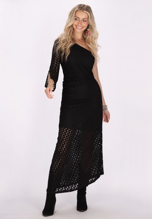Schwarzes schulterfreies Kleid aus strukturiertem Stoff mit gemusterten Ausschnitten, bis zu den Knöcheln fließend, kombiniert mit knielangen schwarzen Stiefeln.