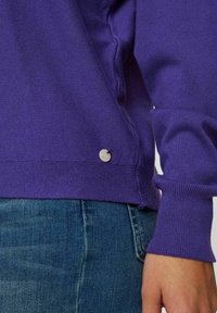 Lila langärmliges Hemd mit gerippten Bündchen und Saum. Verfügt über eine kleine, runde, hellfarbene Akzenttaste an der Seitennaht.