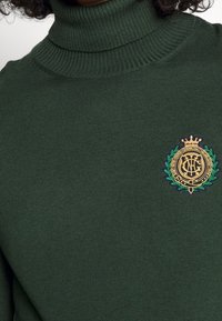 Dunkelgrüner Rollkragenpullover aus geripptem Stoff mit einem goldenen gestickten Logo mit einer Krone und dekorativen Lorbeerblättern.