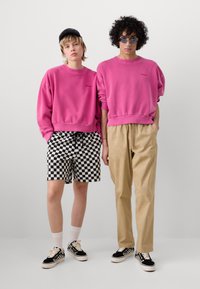 Två personer står sida vid sida, iklädda rosa sweatshirts. Den ena har rutiga shorts, medan den andra har ljusbeige byxor. Båda har sneakers.