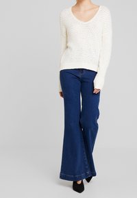 Kremowy sweter z dzianiny o teksturowanym wzorze i szerokimi rękawami, w zestawieniu z luźnymi, ciemnoniebieskimi dżinsami w kształcie dzwonów i czarnymi szpilkami.