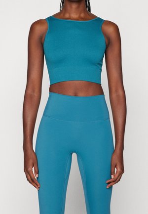 Vrouw draagt een teal mouwloos crop top en teal leggings met hoge taille tegen een effen lichte achtergrond.