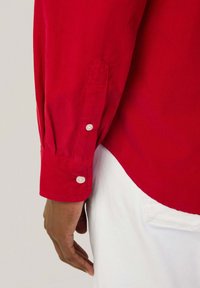 Camisa de corduroy rojo de manga larga con puños abotonados, que presenta una textura suave y un dobladillo redondeado, exhibida sobre un fondo claro.