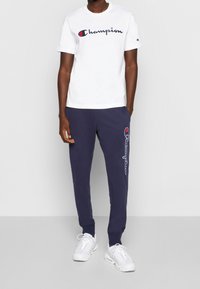 Bílé bavlněné tričko s logem "Champion" v námořnické modři a červené; námořnické modré tepláky nesou logo na levé noze; doplněno bílými sportovními botami.