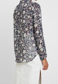 Långärmad blus i genomskinligt marinblått tyg med blommönster i ljusrosa och vitt. Snygg böjd fåll och knappdetalj bak.