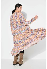 Lang, vloeiende jurk met een ingewikkeld geometrisch patroon in oranje, paars en teal op een witte achtergrond. Lange mouwen met manchetdetails.