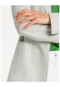 Blazer rayé en vert clair et blanc, avec un col cranté, une poche unique et un tissu lisse à rayures verticales.