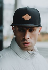 Mustapohjainen snapback-lippis, jossa on ruskea nahkalaatta, johon on painettu "BLACKSKIES"-logo. Lippis on musta, rakenteellinen muotoilu ja litteä lippa.