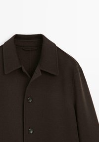 Cappotto di lana marrone scuro con colletto classico, chiusura frontale a bottoni e una finitura in tessuto testurizzato. I bottoni sono neri e rotondi.