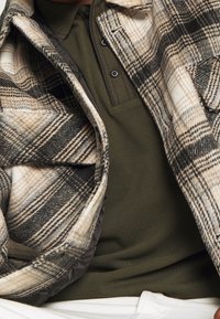 Rutigt flanelljacka i beige, svart och vitt över en mörkgrön polo. Har en knappad krage och mjuk textur.