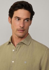 Camicia di lino verde oliva chiaro con colletto button-down, bottoni bianchi e un piccolo logo blu ricamato sul lato sinistro del petto.