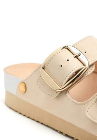 Sandalia beige con hebilla dorada, material de ante, diseño de punta abierta y una suela texturizada sutil. Presenta un acento blanco elegante.