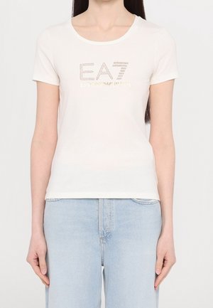 Kobieta ubrana w białą koszulkę z krótkim rękawem z napisem „EA7 EMPORIO ARMANI” z kamieniami sztucznymi, w połączeniu z jasnoniebieskimi dżinsami.