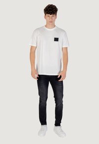 Camiseta de algodón blanca con un bolsillo cuadrado negro, combinada con jeans de mezclilla oscura y zapatillas blancas. Ajuste simple y casual.