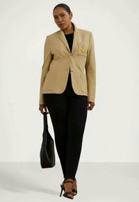 Sacou bej cu croială ajustată, închidere cu buton auriu și un buzunar pe piept brodat cu un logo. Asortat cu pantaloni negri și o geantă neagră.