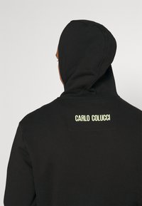 Czarna bluza z kapturem, z prostokątną metką na plecach, na której widnieje napis "CARLO COLUCCI" w jasnooliwkowym kolorze. Miękka faktura materiału.