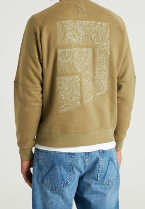 Rückansicht einer Person, die einen hellbraunen Pullover mit abstraktem weißem Stickmuster und blaue Jeans trägt.