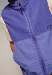 Chaleco de forro polar y nylon púrpura de Adidas con cremallera completa, usado sobre una camiseta de manga corta beige, en una persona.
