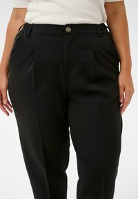 Persoană îmbrăcată cu pantaloni plisati negri, cu un nasture și buzunare laterale, asortați cu un top alb din tricot cu mâneci scurte.