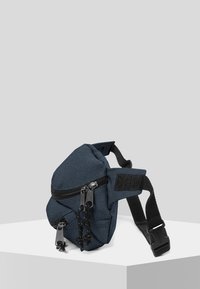 Blå tygaxelväska med triangulär form, utrustad med dragkedjestängning, justerbar rem och texturerade hårdvarudetaljer.