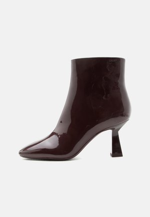 Glinsterende donkerbruine lakleren enkellaars met puntige neus en middelhoge blokhak op effen witte achtergrond.