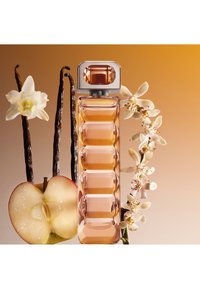 Wysoka, prostokątna butelka perfum z brzoskwiniowym płynem, otoczona strąkami wanilii, białymi kwiatami i połówką jabłka.