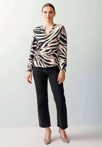 Frau steht vor einer hellen Wand und trägt eine langärmlige Bluse mit Zebra-Streifen, schwarze Jeans und beige hohe Schuhe. Ihr Haar ist zurückgebunden.