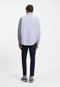 Mężczyzna w jasnoniebieskiej koszuli z długim rękawem, granatowych dopasowanych spodniach i niebieskich tenisówkach, stojący tyłem na białym tle.