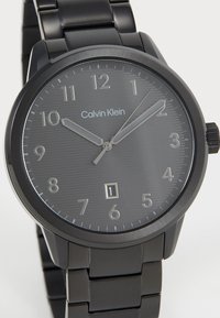 Montre en acier inoxydable noir avec un cadran gris texturé, des chiffres arabes argentés et un affichage de la date. Elle possède un bracelet en métal et un design épuré.