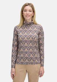 Frau mit glattem, blondem Haar, die einen gemusterten, langärmeligen Rollkragenpullover und beige Hosen trägt, steht vor einem einfarbigen Hintergrund.