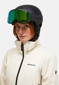 En ung person med fregner iført en sort skihjelm, grønne briller og en cremefarvet PeakPerformance-jakke med høj krave.