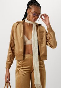 Beiger Velours-Trainingsanzug, bestehend aus einer Reißverschlussjacke mit Kapuze und gesticktem Logo, kombiniert mit passenden Hosen und einem Spitzenschal. Transparente Sonnenbrille.