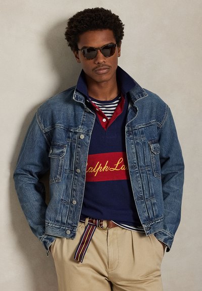 Traper jakna svijetloplave boje s vidljivim šavovima. Ispod je slojeviti navy polo s crvenim detaljima i prugama. Bež hlače sa prugastim remenom.