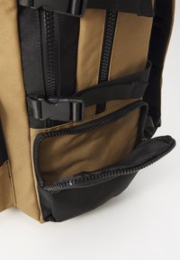 Backpack featuring a tan and black color scheme, durable fabric, multiple zippers, adjustable straps, and a front zippered pocket.