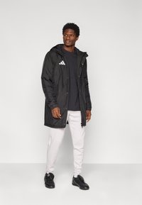 Manteau noir matelassé avec une capuche, fermeture éclair, et trois bandes blanches sur le côté gauche de la poitrine. Associé à une chemise sombre et un pantalon clair.